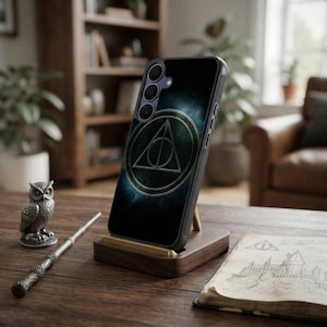 May include: A black smartphone case featuring a circular Deathly Hallows symbol in metallic silver tones against a dark, textured background. The case has a textured side grip and is positioned upright on a wooden stand on a dark surface.
