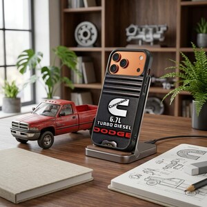 Pode incluir: Uma capa de smartphone com design preto canelado, o logótipo Cummins, o texto 6.7L TURBO DIESEL e a palavra DODGE a vermelho. A capa está montada num suporte de carregamento metálico e apresenta um módulo de câmara laranja na parte traseira.