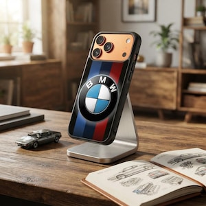 以下が含まれることがあります： 青、紫、赤の縦縞模様の中央にBMWのロゴが配置されたスマートフォンケース。ケースには銅色のカメラハウジングがあり、銀色の金属製スタンドに立てかけられています。背面には3つのカメラレンズが備わっています。