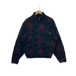 May include: A long-sleeve fleece jacket featuring a dark teal and maroon geometric pattern. The jacket has a full-length front zipper, a high mock neck collar, and a single horizontal zippered chest pocket on the left side, all hanging on a hanger.