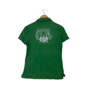 May include: A green short-sleeved polo shirt featuring a collar and a white graphic print on the back. The design includes a shield emblem with mountain shapes, a circular sun, and the text "ELLEN JUNGLE BEACH" surrounded by ornate scrollwork.