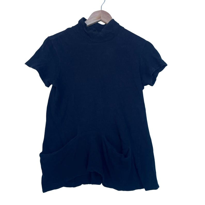May include: A solid black short-sleeved top featuring a mock neck collar and two large patch pockets positioned at the lower front. The garment is displayed on a wooden hanger and is constructed from a soft, textured fabric with a relaxed fit.