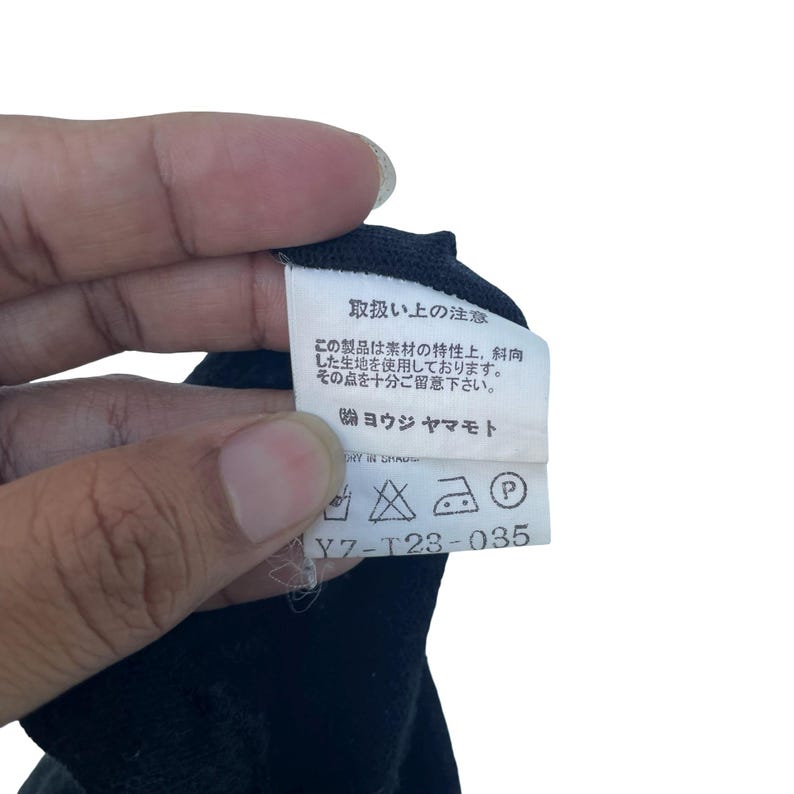 May include: A white rectangular garment care label attached to a black fabric item. The label features black Japanese text, the brand name Yohji Yamamoto, and a series of five laundry care symbols, along with the alphanumeric code Y7-T23-035.