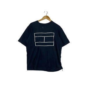Peut inclure: Un t-shirt à manches courtes bleu marine foncé avec un contour graphique rectangulaire blanc divisé en trois sections sur la poitrine. Le vêtement est présenté sur un cintre en bois, avec un col rond et un ourlet droit standard.