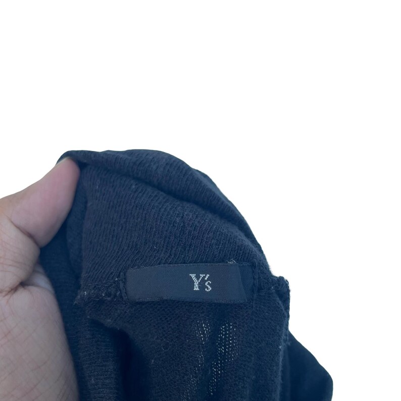 May include: A black knit garment featuring a rectangular fabric label sewn onto the interior neckline. The label is black with the text Y's printed in a white, sans-serif font. The fabric has a visible, fine-gauge knit texture throughout.