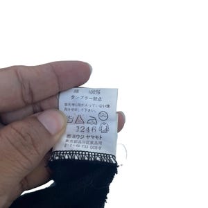 May include: A white garment care label with black text and symbols, indicating 100% cotton composition. The label features Japanese instructions, laundry care icons, the number 1246, and the company name Yohji Yamamoto with a Tokyo address.