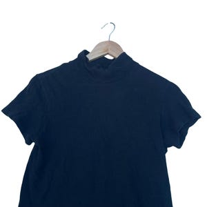 May include: A solid black short-sleeved shirt featuring a mock neck collar. The garment is displayed on a light-coloured wooden hanger, showcasing a simple design with a high neckline and short sleeves, constructed from a plain, dark-coloured fabric.
