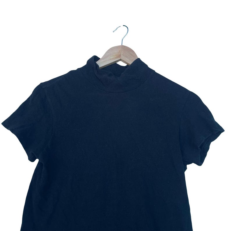 May include: A solid black short-sleeved shirt featuring a mock neck collar. The garment is displayed on a light-coloured wooden hanger, showcasing a simple design with a high neckline and short sleeves, constructed from a plain, dark-coloured fabric.