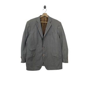 May include: A light gray blazer featuring dark vertical pinstripes, a notched lapel, two front buttons, and two flap pockets. The interior is lined with a brown fabric, and the sleeves are finished with small buttons at the cuffs.