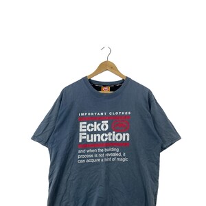 May include: A blue short-sleeve t-shirt featuring the white and red text "IMPORTANT CLOTHES Ecko Function" and the phrase "and when the building process is not revealed, it can acquire a hint of magic" printed on the front chest area.