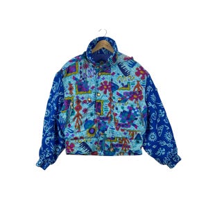 May include: A multi-colored jacket featuring a light blue base with a vibrant, abstract pattern in shades of pink, purple, and dark blue. The sleeves are solid blue with white graphic symbols. It has a high collar, gold-tone buttons, and zippers.