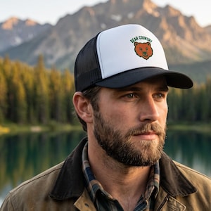 May include: A trucker cap featuring a white front panel with a brown bear illustration and the text BEAR COUNTRY in green letters. The cap has a black brim and black mesh side and back panels, designed with a structured crown and a snapback closure.