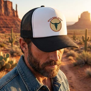 Peut inclure: Une casquette de type trucker noire et blanche avec un motif de crâne de taureau vert foncé dans un cercle beige bordé d'une corde. Le texte "WILD WEST SPIRIT" est inscrit en arc de cercle au-dessus du crâne en brun. Visière et filet noirs.