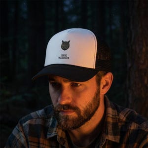 May include: A trucker hat with a white front panel, a black brim, and black mesh back panels. The front features a dark gray graphic of an owl head centered above the black text "NIGHT WANDERER" in a clean, sans-serif font style.