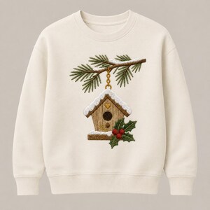 May include: A cream-coloured crewneck sweatshirt featuring a central embroidered design of a wooden birdhouse hanging from a pine branch. The birdhouse is topped with white snow, a small gold heart, and is decorated with green holly and red berries.