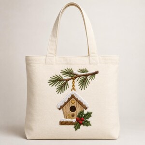 May include: A cream-coloured canvas tote bag featuring an embroidered design of a wooden birdhouse hanging from a snow-dusted pine branch. The birdhouse has a small gold heart detail, and the scene is accented with green holly leaves and red berries.