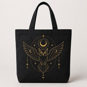 May include: A black tote bag featuring a gold-colored graphic print of an owl with outstretched wings. The design includes a crescent moon, radiating sunburst lines, and various star shapes arranged in a circular pattern around the owl.