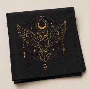 May include: A square black fabric napkin featuring a gold embroidered design of an owl with outstretched wings. The owl is centered within a circular frame of stars and crescent moons, with additional hanging star and moon motifs below.