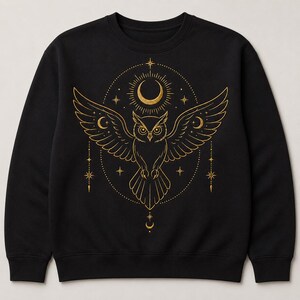May include: A black long-sleeve sweatshirt featuring a gold-colored graphic print of an owl with outstretched wings. The design includes a crescent moon, stars, and celestial elements arranged in a circular pattern centered on the chest area.