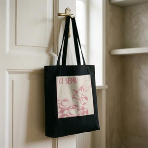 Puede incluir: Un bolso tote negro con un estampado gráfico cuadrado en tonos rosados. El diseño muestra una mano vertiendo café de una cafetera italiana en una taza sobre un mantel de cuadros, con el texto CI SEMU impreso en una fuente sans-serif.