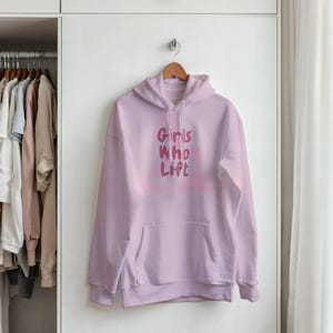 Puede incluir: Una sudadera con capucha de color rosa claro con mangas largas, un bolsillo tipo canguro en la parte delantera y el texto "Girls Who Lift" impreso en un tono rosa más oscuro en el pecho. La prenda cuelga de una percha de madera en una puerta blanca.