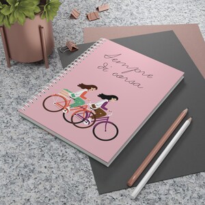 Puede incluir: Un cuaderno de espiral con una cubierta de color rosa claro que presenta una ilustración gráfica de dos personas montando en bicicleta. El texto "Sempre di corsa" está impreso en letra cursiva negra en la cubierta.