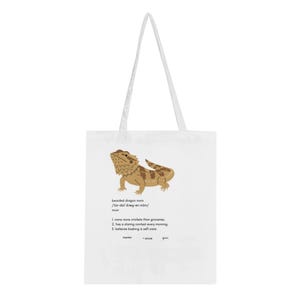 Puede incluir: Una bolsa de tela blanca con una ilustración de un dragón barbudo marrón sobre un texto estilo diccionario que define bearded dragon mom, incluyendo tres puntos numerados sobre grillos, concursos de miradas y tomar el sol, con líneas en blanco.
