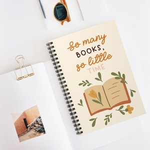 Puede incluir: Un cuaderno de espiral con cubierta color crema que presenta el texto "So many BOOKS, so little TIME" en marrón y rosa, acompañado de un gráfico de un libro abierto con una flor amarilla y hojas verdes, además de un clip dorado y unas gafas de sol.