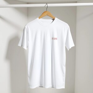 Puede incluir: Una camiseta blanca de manga corta colgada en una percha de madera. La prenda presenta un pequeño parche rectangular en la parte superior izquierda del pecho con el texto CI SEMU impreso en rojo oscuro sobre un fondo rosa claro.