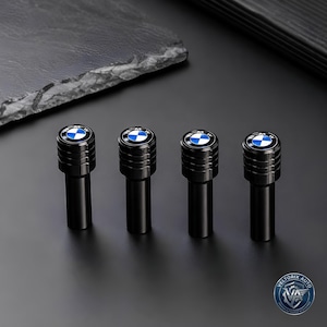 May include: A set of four black cylindrical tire valve stem caps featuring a grooved top section and a circular BMW logo emblem with blue, white, and black colors on the top surface. The caps are arranged in a row against a dark background.