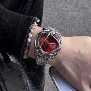 Könnte beinhalten: Eine silberfarbene Armbanduhr mit einem leuchtend roten Zifferblatt, das in einen komplexen, organisch geformten Metallrahmen eingefasst ist. Die Uhr verfügt über ein Metallgliederarmband und eine Datumsanzeige sowie ein Perlenarmband.