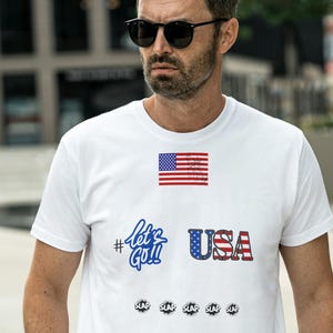 以下が含まれることがあります： アメリカ国旗のグラフィック、#Let's Go!というテキスト、国旗柄のUSAというテキスト、そして5つの小さな黒い円形アイコンが並んだ白い半袖Tシャツ。クルーネックでスタンダードなフィット感のTシャツです。