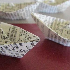 May include: A collection of small paper boats made from pages of a book. The boats are folded in a traditional origami style and are arranged on a red surface. The text on the pages of the book is visible on the boats.