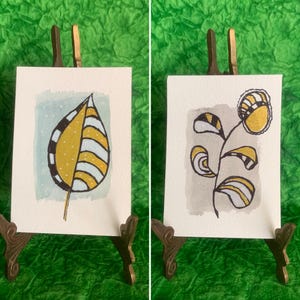 May include: Two small watercolor paintings on miniature easels. One features a yellow and white leaf design with black outlines. The other depicts a yellow and white flower with black outlines. Both are on white paper.