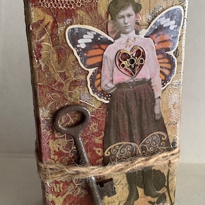 May include: A mixed media art piece featuring a vintage image of a woman with butterfly wings and a heart-shaped embellishment. The artwork is adorned with a rusty key and wrapped with twine. The background has a textured, patterned design in gold and burgundy.