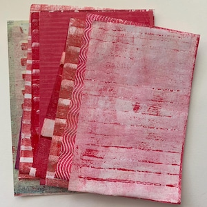 May include: A stack of handmade paper sheets in varying shades of red, with white and red patterns. The papers have textured surfaces with printed designs, including stripes, wavy lines, and block patterns. The papers are arranged on a white surface.