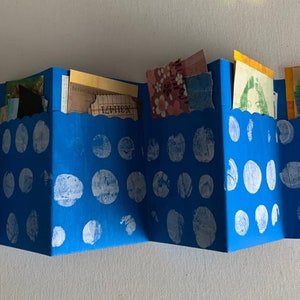 May include: Accordion-style blue paper art piece with white polka dots. The piece is filled with various colorful paper scraps and small cutouts. The piece is held together with a small metal clasp.