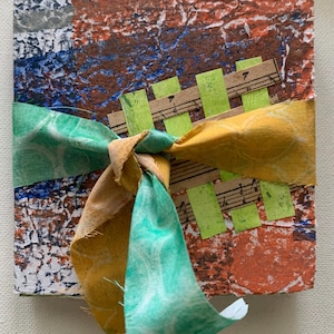 May include: A handmade journal with a textured, colorful cover in shades of red, blue, and white. The journal is tied with a green and yellow patterned ribbon. Decorative elements include musical notes and green paper accents.