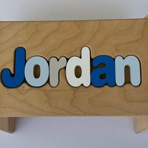 May include: Wooden step stool with the name "Jordan" spelled out in blue, light blue, gray, and white letters. The stool is made of light-colored wood and has a simple, modern design.