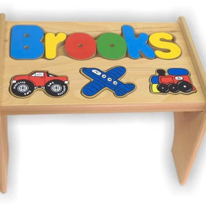 May include: Wooden children's table with the name "Brooks" spelled out in colorful, raised letters. The table features a red monster truck, a blue airplane, and a red and yellow train. The table is made of light-colored wood.