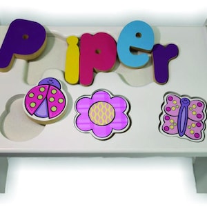 May include: A white wooden puzzle board with the name "Piper" spelled out in colorful, wooden letters. The board also features wooden cutouts of a ladybug, a flower, and a butterfly.
