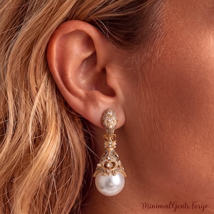 May include: A gold-toned drop earring featuring an ornate, filigree-style top section encrusted with small clear stones and dark accents. A large, smooth, spherical white pearl hangs from the base of the intricate metalwork, creating a long silhouette.