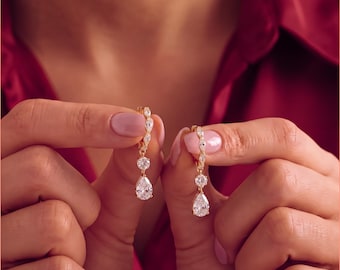 May include: A pair of gold-toned drop earrings featuring a small hoop at the top, a central round clear stone, and a dangling pear-shaped clear stone at the bottom. The earrings are held up to display the clear, faceted stones and the metallic gold finish.