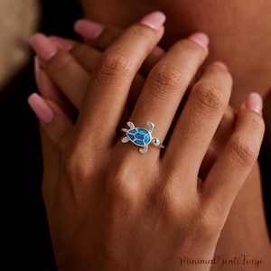 Puede incluir: Un anillo de plata con un diseño de tortuga marina con una incrustación de ópalo azul en el caparazón. La cabeza y las aletas de la tortuga están decoradas con pequeñas piedras transparentes. El aro es fino y de plata pulida.