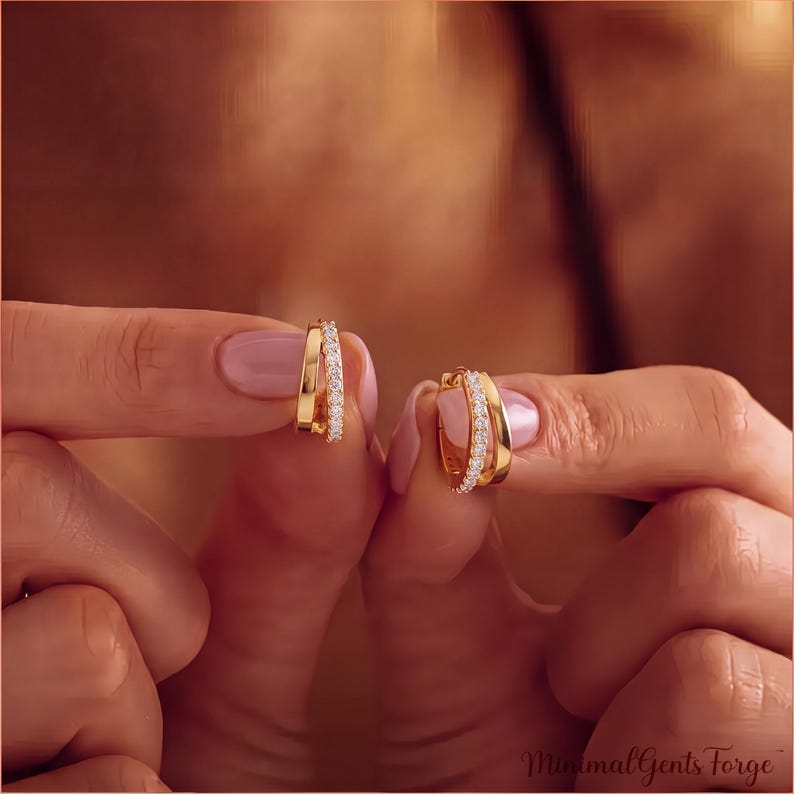 May include: A pair of gold hoop earrings, each featuring a polished gold band paired with a thinner band encrusted with small, clear, sparkling stones. The earrings are held up to display the dual-band design and the reflective metallic finish.