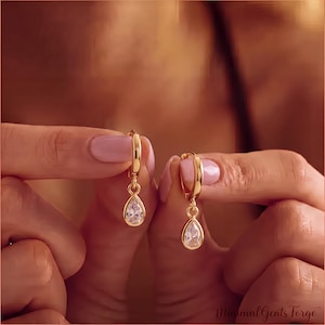 May include: A pair of gold hoop earrings featuring a dangling teardrop-shaped clear gemstone. Each earring consists of a polished gold circular hoop connected to a single pear-cut white stone, creating a simple and elegant jewelry design.
