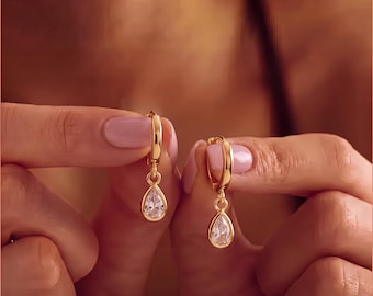 May include: A pair of gold hoop earrings featuring a dangling teardrop-shaped clear gemstone. Each earring consists of a polished gold circular hoop connected to a single pear-cut white stone, creating a simple and elegant jewelry design.