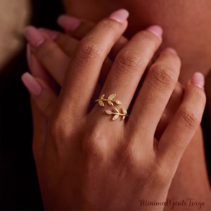 May include: A gold-toned open ring featuring two delicate, overlapping leaf-shaped branches. Each leaf is accented with small, clear, sparkling stones. The ring has a bypass design, leaving a small gap between the two leaf-tipped ends.