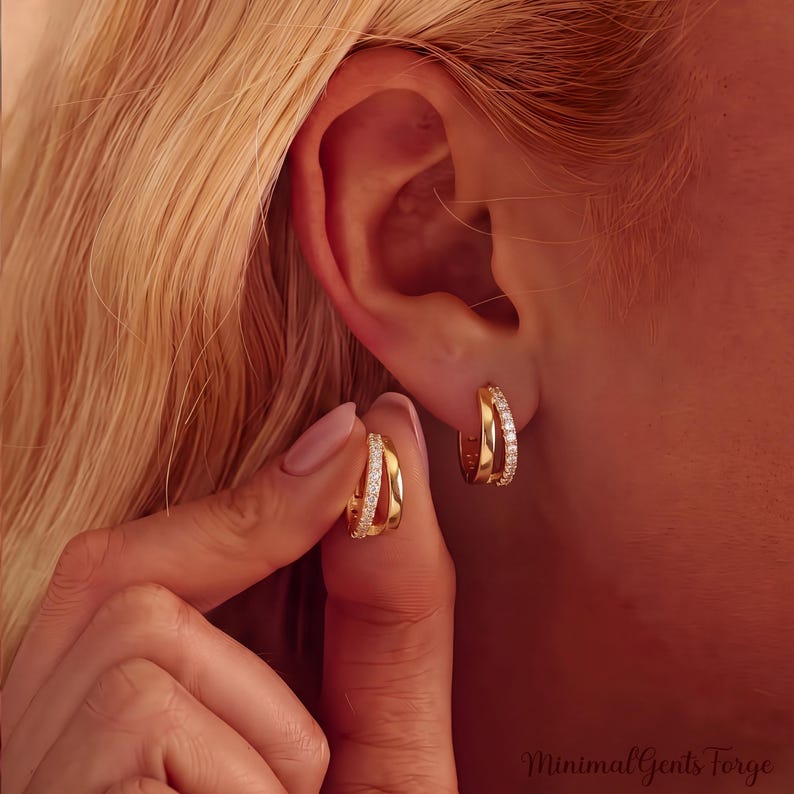 May include: A pair of gold hoop earrings featuring a dual-band design. One band is polished gold, while the other is encrusted with small, clear, sparkling stones. The earrings are small, circular, and have a hinged closure mechanism.