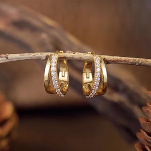 May include: A pair of gold hoop earrings featuring a double-band design. One band is smooth and polished gold, while the other is encrusted with a row of small, clear, sparkling stones. The earrings are hinged and have a secure clasp mechanism.